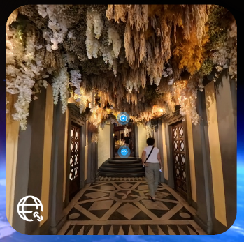 A woman walking down a hallway with a geometic-designed floor, with cream-colored plants hanging down from and covering the ceiling.
