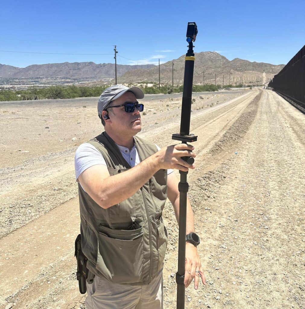 A man wearing black sunglasses and gray vest holds a black tripod 